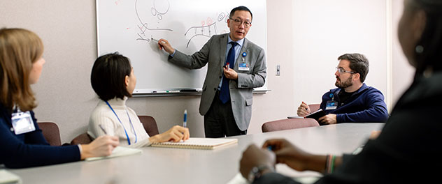 An investigator points to a white board while colleagues observe.