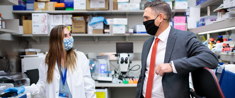 Image showing Nathan K. LeBrasseur, Ph.D., M.S., talking with a lab worker in a white coat, both wearing masks