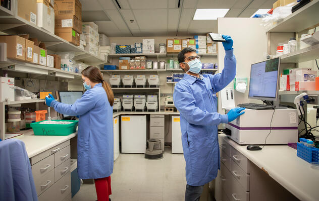 Photograph showing two gowned and masked lab workers standing at tables and managing samples